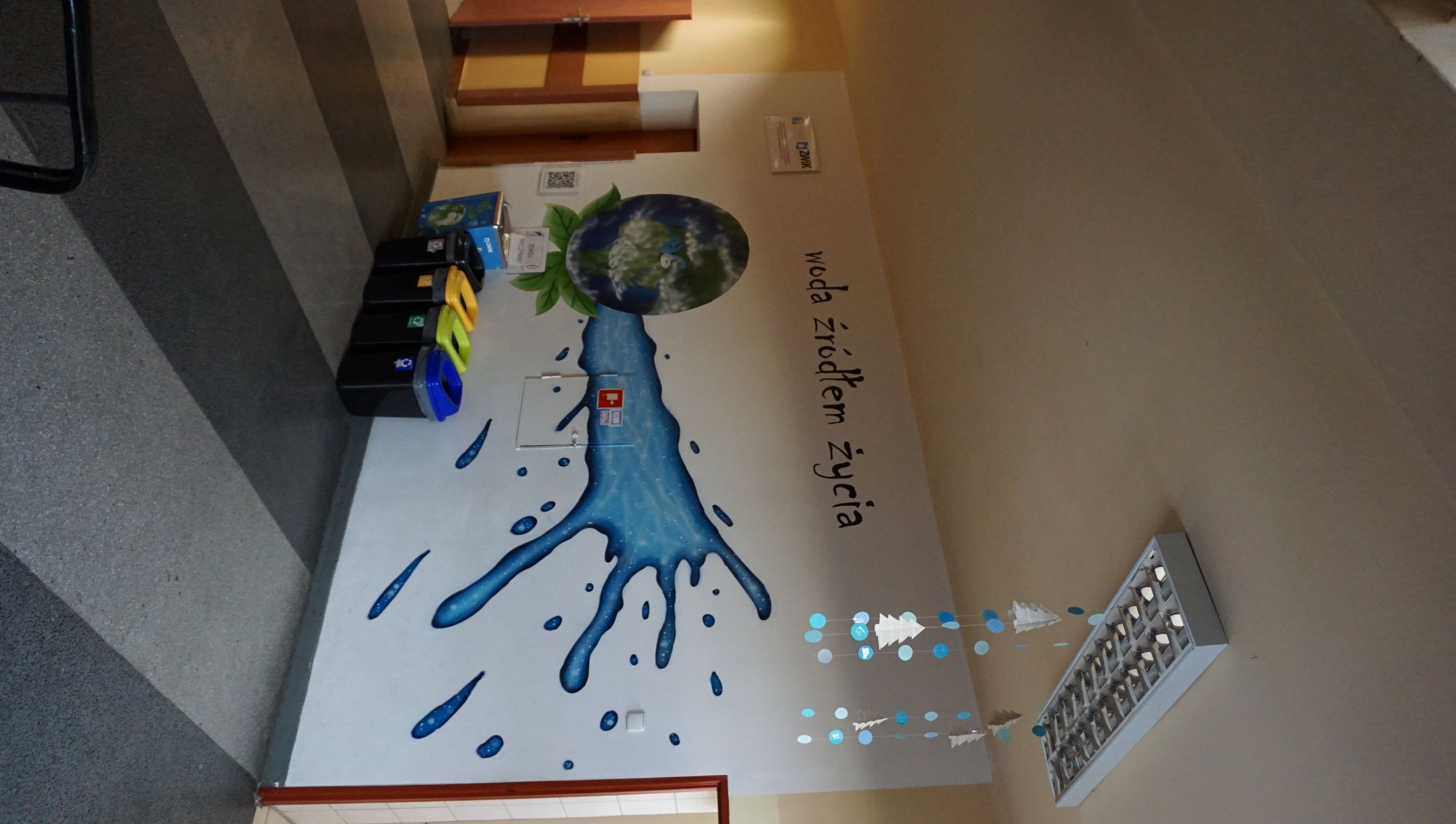 Fragment szkolnego korytarza z dużym, kolorowym muralem na ścianie przedstawiającym niebieski strumień wody tryskający z planety Ziemia, otoczonej zielonymi liśćmi. Nad grafiką widnieje czarny napis: &bdquo;woda źr&oacute;dłem życia&rdquo;. Pod malowidłem znajduje się zestaw czterech koszy do segregacji odpad&oacute;w: czarny, ż&oacute;łty, zielony i niebieski, każdy oznaczony odpowiednim symbolem. Na suficie wiszą papierowe ozdoby w kształcie choinek i kulek. Na ścianie obok drzwi widoczny jest logotyp ZWiK oraz skrzynka przeciwpożarowa. Posadzka ma szare i czarne pasy.