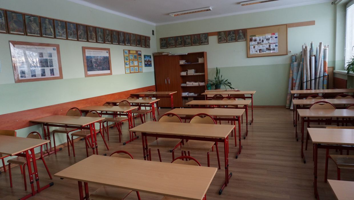 Sala historyczna. Sala lekcyjna w szkole z jasnymi, zielono-białymi ścianami. W pomieszczeniu znajdują się rzędy dwuosobowych ławek i krzeseł z czerwonymi metalowymi stelażami oraz jasnobrązowymi blatami i oparciami. Po lewej stronie widać plansze edukacyjne i obrazy historyczne w ramach, zawieszone w dw&oacute;ch rzędach na ścianie. Z tyłu klasy znajduje się drewniana szafa oraz regał z ułożonymi stosami książek. Po prawej stronie, przy oknie, stoją zwinięte mapy w tubach oraz doniczki z roślinami. Podłoga pokryta jest panelami imitującymi drewno, a przez okno wpada dzienne światło.