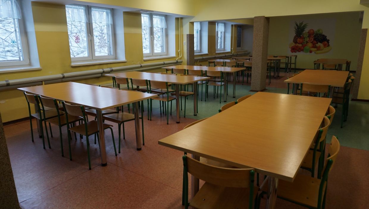 Stoł&oacute;wka. Szkolna stoł&oacute;wka z jasnymi ż&oacute;łtymi ścianami i wieloma prostokątnymi stołami otoczonymi drewnianymi krzesłami z metalowymi, zielonymi nogami. W pomieszczeniu jest kilka rzęd&oacute;w okien z firankami, przez kt&oacute;re widać śnieg i gałęzie drzew. Na tylnej ścianie widnieje duża ilustracja przedstawiająca kompozycję owoc&oacute;w, m.in. ananasa, banana i winogron. Pod oknami biegną poziome rury grzewcze, a podłoga składa się z kolorowych segment&oacute;w w odcieniach czerwonym i zielonym.