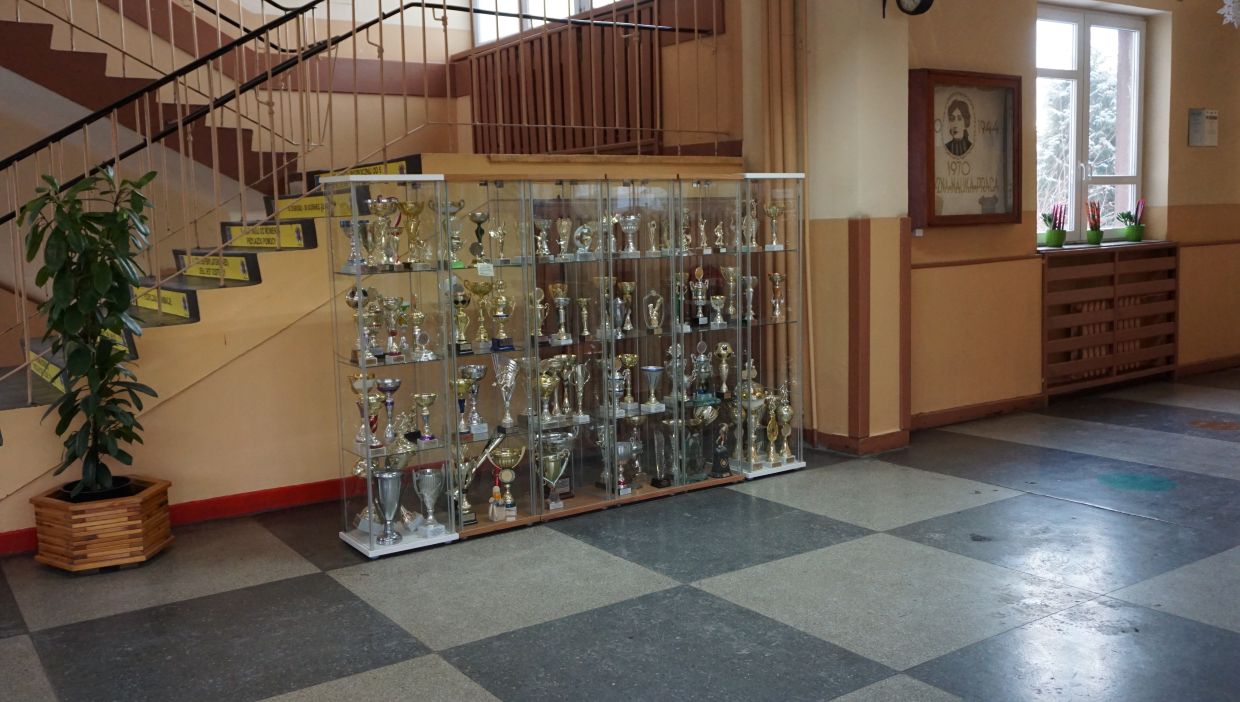 Hol szkoły. Hol szkolny z przeszkloną gablotą pełną puchar&oacute;w sportowych ustawioną przy ścianie pod schodami. Puchary są r&oacute;żnej wielkości i kształtu, wykonane ze srebrnego i złotego metalu. Po lewej stronie stoi duża roślina doniczkowa. W tle klatka schodowa z ż&oacute;łtymi tabliczkami na stopniach oraz okno z doniczkami z kwiatami. Na ścianie widoczny jest portret osoby z napisem zawierającym lata &bdquo;1944&rdquo; i &bdquo;1970&rdquo;.