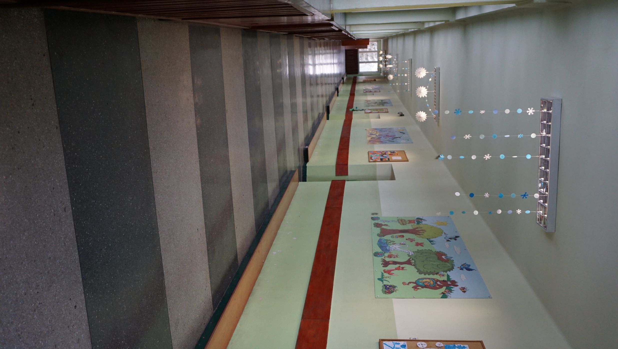 Korytarz drugie piętro. Szkolny korytarz z jasnozielonymi ścianami, ozdobiony kolorowymi rysunkami i tablicami edukacyjnymi. Na suficie zawieszone są zimowe ozdoby, m.in. papierowe śnieżynki oraz girlandy z niebieskimi i białymi krążkami. Pod ściennymi obrazkami biegnie drewniana listwa ochronna. Posadzka ma naprzemienne pasy w kolorze szarym i grafitowym. Po lewej stronie znajdują się okna z widokiem na drzewa, a poniżej nich &mdash; drewniane osłony kaloryfer&oacute;w.