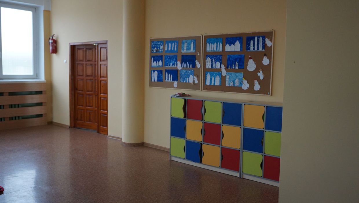 Sala klasy pierwszej. Sala z ż&oacute;łtymi ścianami, na kt&oacute;rych znajdują się dwie tablice korkowe z pracami plastycznymi przedstawiającymi zimowe krajobrazy z domkami i bałwankami. Poniżej znajduje się rząd kolorowych szafek uczniowskich w kolorach: czerwonym, ż&oacute;łtym, zielonym i niebieskim. Po lewej stronie widoczne są drewniane drzwi oraz okno z parapetem i grzejnikiem osłoniętym drewnianą osłoną.