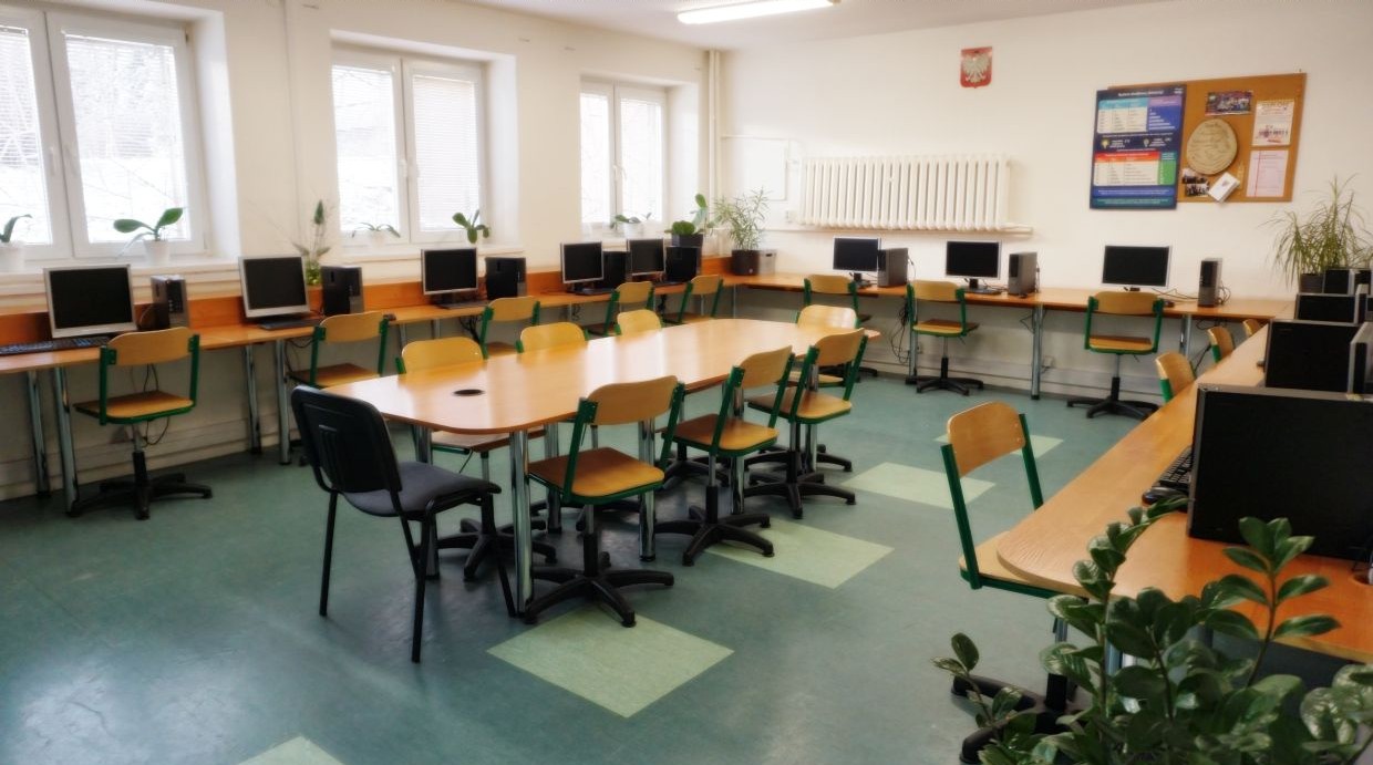 Sala informatyczna. Sala komputerowa w szkole z jasnymi ścianami i zieloną wykładziną podłogową. Wzdłuż ścian ustawione są biurka z komputerami stacjonarnymi oraz krzesła z zielonymi oparciami. Na środku pomieszczenia znajduje się duży, owalny st&oacute;ł z krzesłami do pracy grupowej. Pomieszczenie jest dobrze oświetlone naturalnym światłem wpadającym przez trzy okna, na kt&oacute;rych parapetach stoją doniczki z roślinami. Na ścianie wisi tablica korkowa z informacjami oraz plakat edukacyjny. W pomieszczeniu znajduje się także kilka większych roślin w donicach.