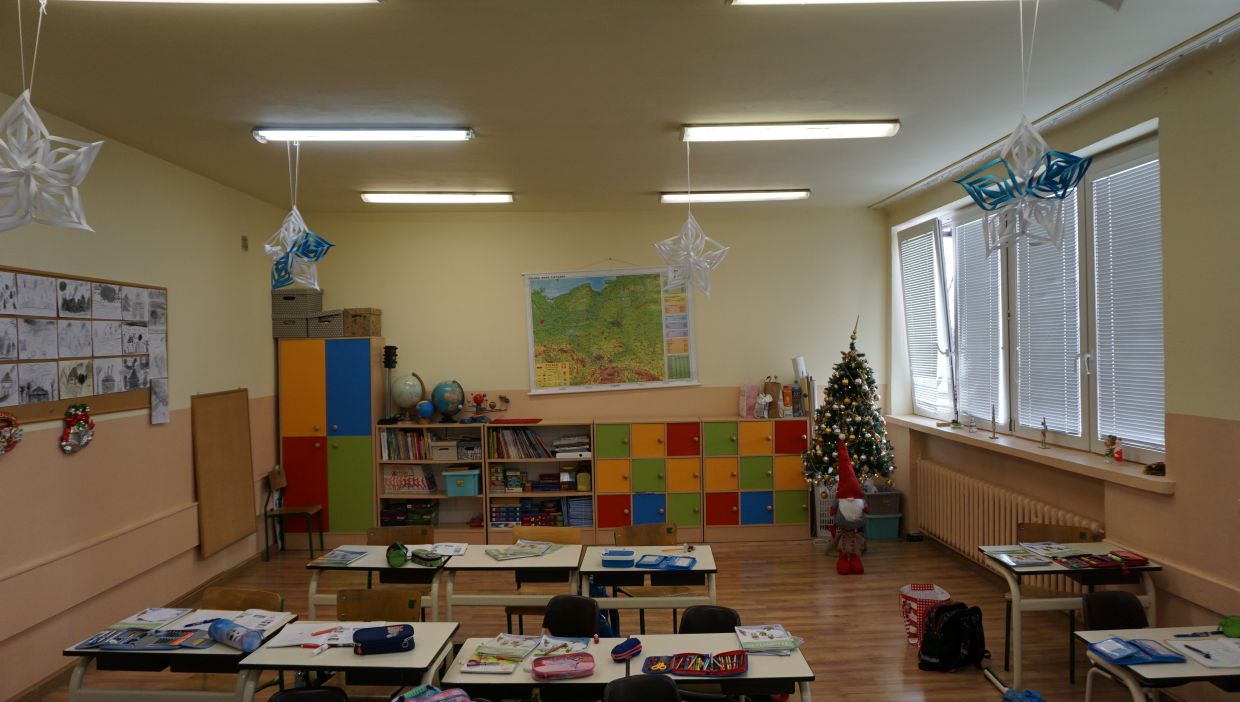 Sala klasy drugiej. Sala lekcyjna z jasnymi ścianami, ozdobiona papierowymi śnieżynkami wiszącymi z sufitu oraz świąteczną choinką ustawioną przy oknach z żaluzjami. Na ścianie wisi mapa Polski. Pod nią i na bokach znajdują się regały z książkami, pomocami naukowymi i kolorowymi szafkami. Na środku sali stoją uczniowskie ławki z rozłożonymi przyborami i zeszytami. Obok choinki stoi duża dekoracja w kształcie skrzata.