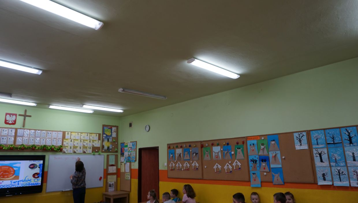 Sala klasy trzeciej. Sala lekcyjna z jasnozielonymi ścianami, w kt&oacute;rej nauczycielka pisze na białej tablicy, a grupa uczni&oacute;w siedzi na krzesełkach z boku. Na ścianach wiszą plansze edukacyjne, kolorowe prace plastyczne dzieci oraz tablice z cyframi i literami. Widoczne są także krzyż i godło Polski.