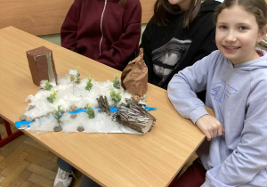 Trzy uczennice prezentują makietę zimowej Narnii z rzeką i ośnieżonym krajobrazem wykonanym z naturalnych materiałów.