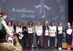 Uczestnicy i organizatorzy akcji Aniołkowe Granie stoją na scenie pod ekranem z logo wydarzenia.