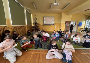 Grupa dzieci siedzi na dywanie w świetlicy i czyta listy, w tle choinka oraz ekran z prezentacją projektu.
