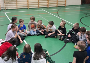 Grupa uczniów siedzi w kręgu na sali gimnastycznej podczas wspólnej zabawy.