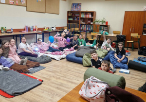 Uczniowie siedzą na materacach i śpiworach w klasie przygotowanej do noclegu.