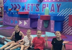 Cztery uczennice odpoczywają na miękkich matach w kolorowej strefie trampolin. W tle widoczne są elementy torów przeszkód oraz dynamiczna, multimedialna przestrzeń sprzyjająca aktywnej zabawie.