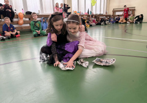 Dwie dziewczynki w przebraniach siedzą na podłodze i zgniatają gazety w trakcie konkursu w sali gimnastycznej.