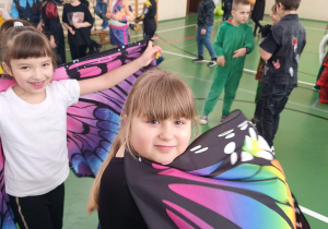 Dwie dziewczynki z kolorowymi skrzydłami motyla stoją w sali gimnastycznej wśród innych przebranych dzieci.