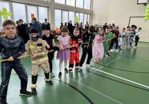 Uczniowie w przebraniach stoją w rzędzie i przeciągają linę, a inne dzieci siedzą na ławkach w tle.