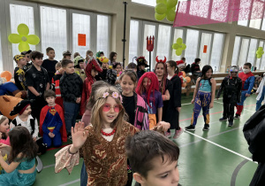 Dziewczynka w kolorowym stroju tańczy w sali gimnastycznej, a za nią stoją inni przebrani uczniowie.