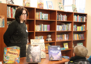 Nauczycielka stoi przed regałem z książkami i prezentuje dzieciom wybrane tytuły.