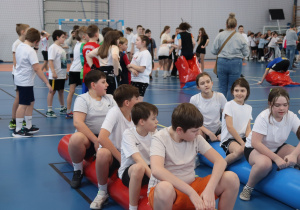 Grupa uczniów siedzi na dmuchanych materacach w hali sportowej podczas wydarzenia.
