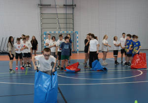 Uczeń biegnie w worku podczas konkurencji sportowej, a w tle stoją inni uczestnicy.