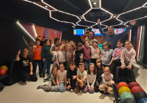 Grupa dzieci wraz z rodzicami pozuje do wspólnego zdjęcia w kręgielni przy torach do kręgli.