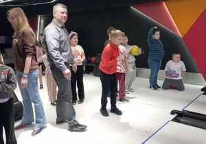 Chłopiec rzuca kulą do kręgli, a inni uczniowie i rodzice stoją w tle przy torze.