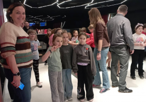 Kilka dziewczynek stoi w kręgielni, a w tle widoczni są rodzice i opiekunowie.