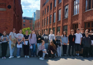 Grupa uczniów pozująca do zdjęcia na zewnątrz przed zabytkowym budynkiem z czerwonej cegły Fabryki Wełny Hotel & SPA, w tle wieża zegarowa i niebo z lekkim zachmurzeniem.