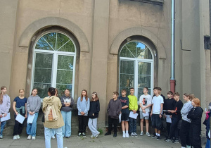 Uczniowie klasy VIIb przed paaUczniowie stoją w półokręgu na tle starego budynku z dużymi oknami, słuchając osoby prowadzącej, która stoi tyłem do aparatu.łacem Kruschego w Pabianiach, w którym obecnie mieści się w nim Urząd Stanu Cywilnego.