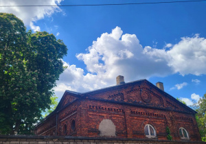 Stare ceglane budynki przemysłowe z czerwonej cegły na tle błękitnego nieba z białymi, puszystymi chmurami. Na pierwszym planie po lewej stronie widać duże, zielone drzewo, a po prawej fragmenty innych drzew. Budynek ozdobiony jest półkolistymi oknami i ceglaną ornamentyką. Całość otoczona jest niskim murem.