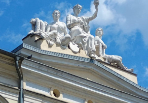 Zdjęcie przedstawia rzeźby trzech klasycznych postaci siedzących na szczycie zabytkowego budynku. Środkowa figura trzyma w górze pochodnię, co może symbolizować wiedzę lub oświecenie. Rzeźby wykonane są w stylu klasycznym, a ich białe sylwetki kontrastują z niebieskim niebem i chmurami w tle.