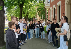 Grupa młodzieży stoi na chodniku w cieniu drzew wzdłuż ulicy, słuchając kobiety przewodniczki. W tle widoczna jest ceglana kamienica z jasnymi obramowaniami okien. Scena odbywa się w słoneczny dzień, a uczniowie trzymają kartki, uczestnicząc w wycieczce edukacyjnej.
