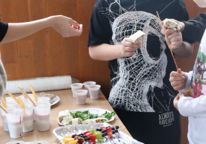 Uczniowie stoją przy stole pełnym zdrowych przekąsek i wybierają jedzenie podczas szkolnego kiermaszu.