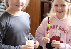 Dwie uczennice trzymają zdrowe przekąski – deser w kubeczku i owocowy szaszłyk podczas szkolnego kiermaszu.