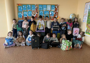 Grupa uczniów siedzi i stoi z różnymi przedmiotami zamiast plecaków, m.in. walizkami i torbami.