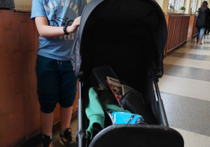 Chłopiec stoi przy wózku dziecięcym, w którym znajdują się książki i przybory szkolne.