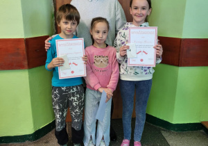 Na zdjęciu stoją cztery osoby: troje dzieci i kobieta (Dyrektorka) na tle drzwi z numerem 28 w szkolnym korytarzu. Dwoje dzieci (chłopiec i dziewczynka) trzymają w rękach dyplomy z napisem „WYRÓŻNIENIE”. Wszyscy uśmiechają się do zdjęcia, a Dyrektorka stoi za dziećmi, obejmując je. Otoczenie ma zielono-białe ściany i szkolną posadzkę.ursie: Grzegorz, zuzanna i Joanna z paną dyrketor Anną Michalak.