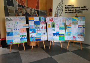 djęcie przedstawia cztery sztalugi ustawione w szkolnym holu przed ścianą z muralem i napisem „Szkoła Podstawowa nr 13 im. Stefanii Sempołowskiej w Pabianicach”. Na sztalugach umieszczone są kolorowe prace plastyczne uczniów klas 1–3, przedstawiające różne sceny i budynki w technice rysunkowej i malarskiej. W tle widoczna jest mozaika ścienna z postaciami oraz duże pióro będące częścią dekoracji ściennej.