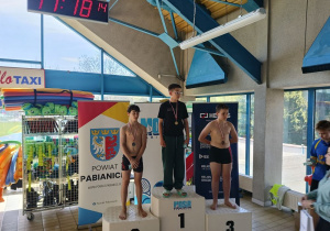 Trzech uczniów stoi na podium i prezentuje medale zdobyte w zawodach pływackich.