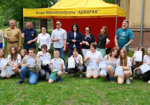 Grupa uczniów i dorosłych stoi razem, trzymając dyplomy po zakończeniu gry miejskiej.