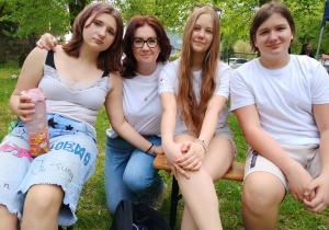 Grupa uczniów i nauczycielka siedzą razem na ławce podczas wydarzenia plenerowego.