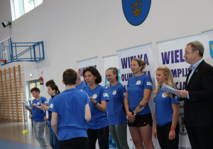 Uczeń odbiera certyfikat od dorosłych podczas wydarzenia sportowego w hali.