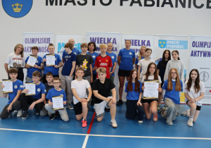 Grupa uczniów i dorosłych stoi w hali sportowej, trzymając certyfikaty uczestnictwa w wydarzeniu sportowym.