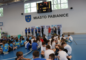 Grupa uczniów siedzi na podłodze hali sportowej podczas spotkania.