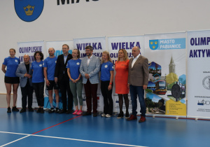 Dorośli uczestnicy wydarzenia stoją obok siebie w hali sportowej podczas oficjalnej części.