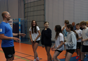 Instruktor prowadzi zajęcia sportowe, a grupa uczniów wykonuje ćwiczenia i rozmawia w hali sportowej.