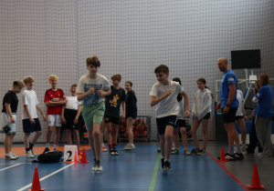 Instruktor rozmawia z grupą uczniów stojących w hali sportowej.