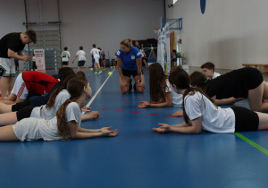 Grupa uczniów ćwiczy w hali sportowej przy sprzęcie treningowym.