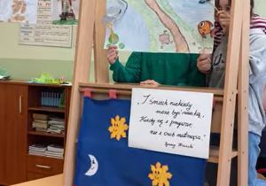 Uczniowie prezentują przedstawienie kukiełkowe przy drewnianej scenie z kolorową ilustracją zwierząt i elementami przyrody.
