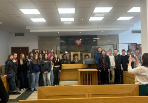 Grupa uczniów pozuje do zdjęcia w sali sądowej. Młodzież stoi przy ławie sędziowskiej, na tle godła Polski umieszczonego nad czarną ścianą z marmuru. Na pierwszym planie widoczne są drewniane ławy dla publiczności. Po prawej stronie kobieta w jasnym swetrze fotografuje grupę. Wnętrze sali jest jasne, z sufitem złożonym z kwadratowych paneli świetlnych.