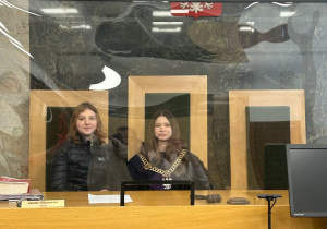 Dwie dziewczyny pozują za stołem sędziowskim w sali rozpraw. Jedna z nich ma na sobie łańcuch sędziowski. Nad nimi widnieje godło Polski.