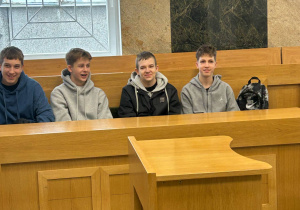 Czterech uczniów siedzi w ławie sali sądowej, za nimi marmurowa ściana i okno z kratą. Wszyscy uśmiechają się do zdjęcia.